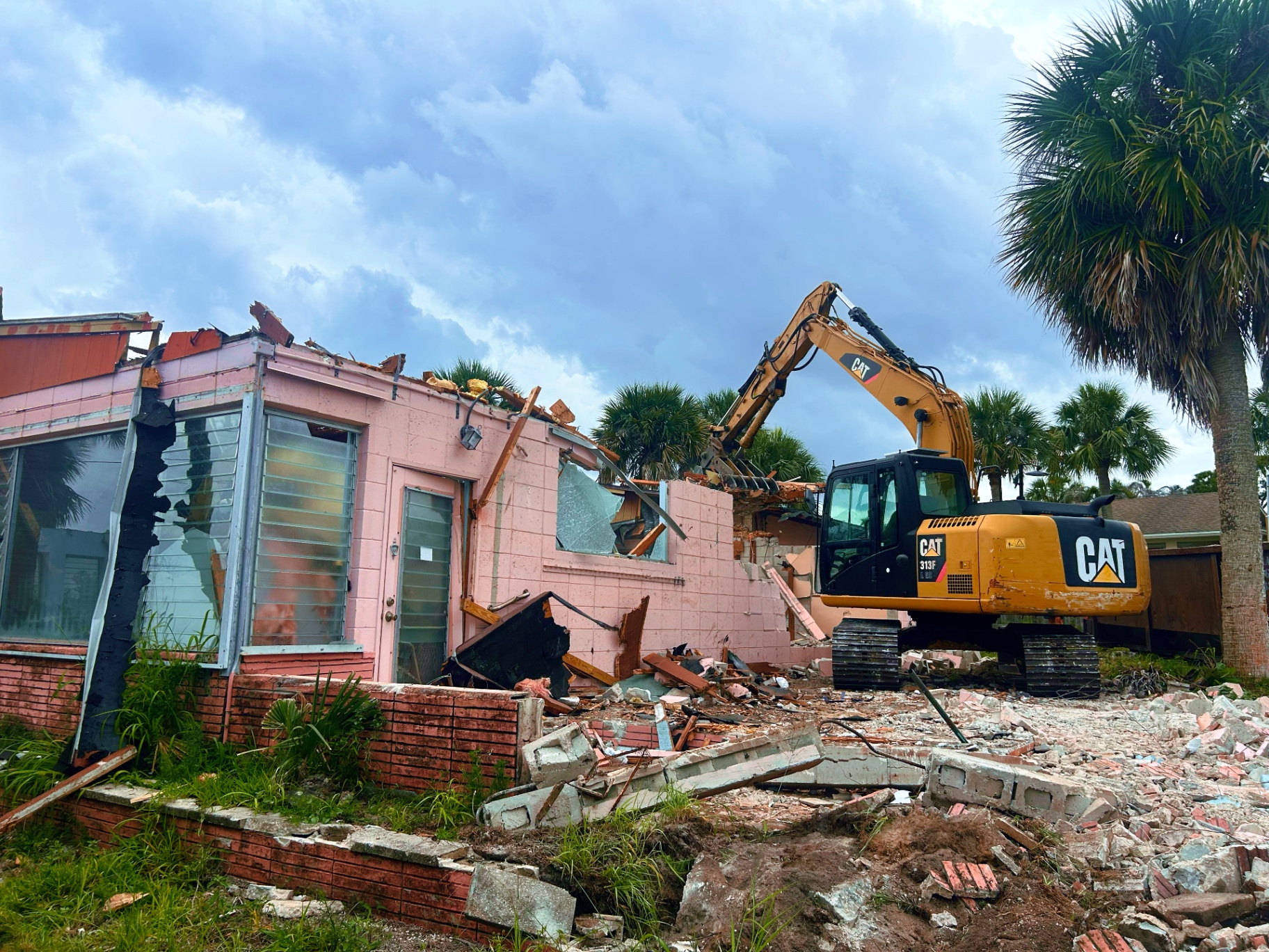 CAT 313F excavator demolishing a pink CBS single-family home in Martin County, FL surrounded by palm trees